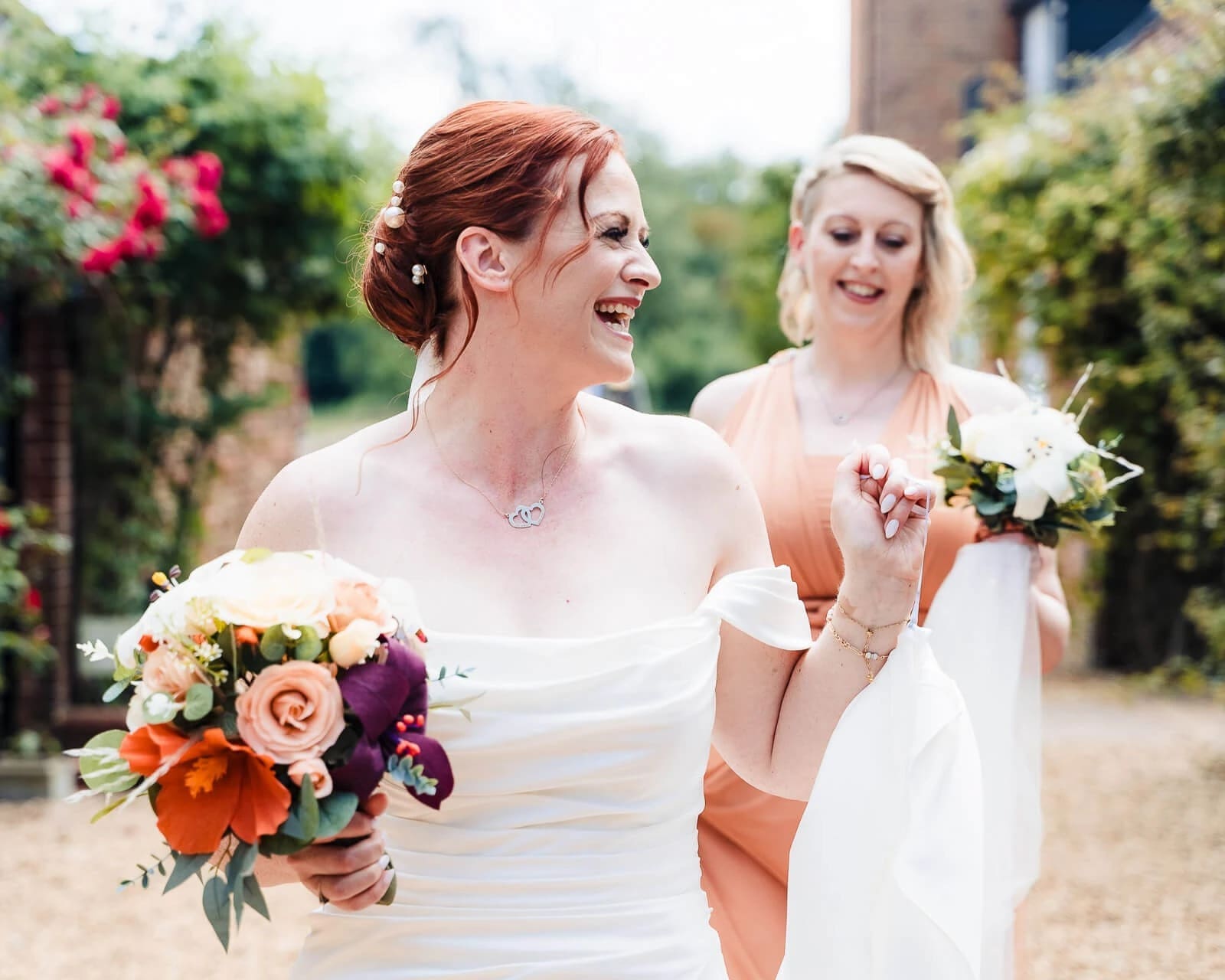 Bride and bridesmaid walking together outdoors, both smiling and holding bouquets. — Wedding at Hunters Hall, Norfolk