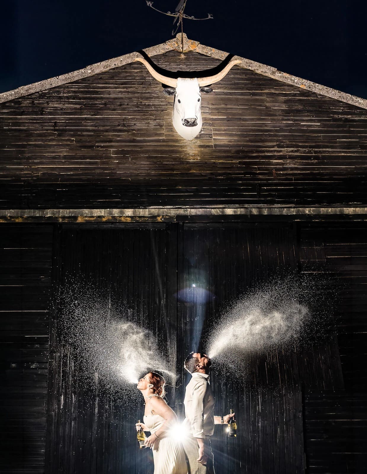 Bride and groom standing together as water sprays arc around them in front of a wooden barn at night. — Wedding at Hunters Hall, Norfolk