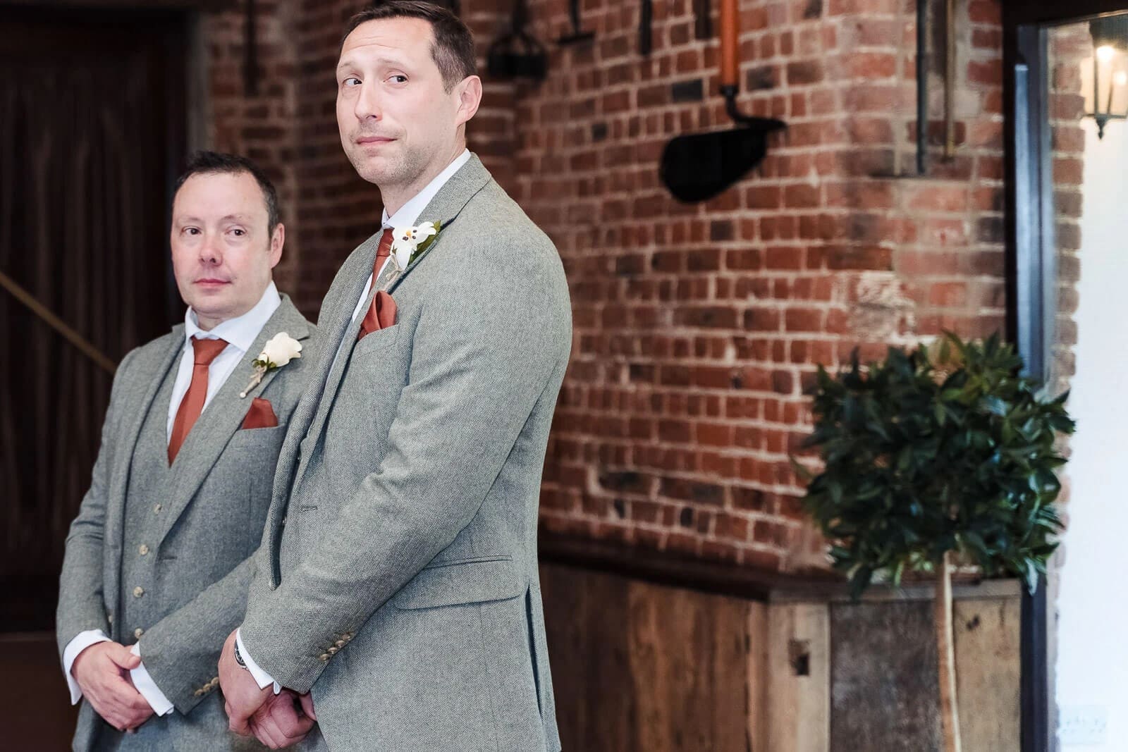 Two men in formal suits stand together at a brick venue, posing for a wedding portrait. — Wedding at Hunters Hall, Norfolk