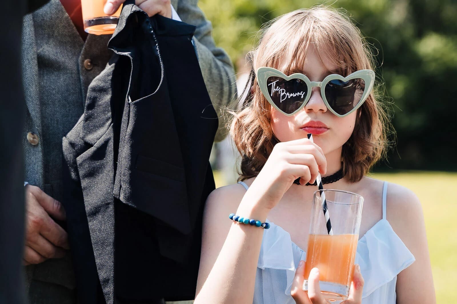 Young girl posing in heart-shaped sunglasses while an adult holds a black garment beside her outdoors. — Wedding at Hunters Hall, Norfolk