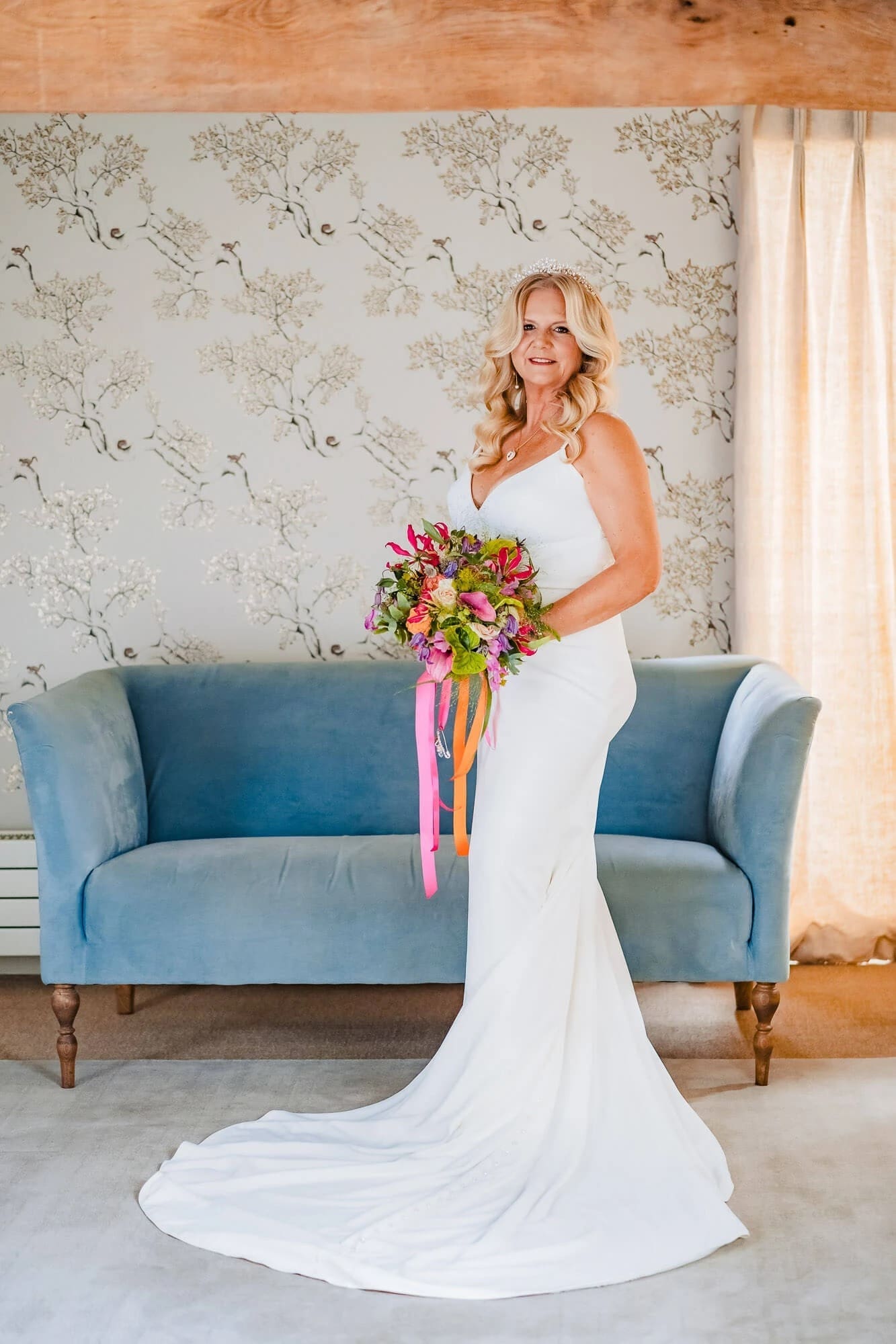 Bride standing with colorful bouquet, posing in front of blue sofa for formal wedding portrait. — Wedding at South Farm, Cambridgeshire