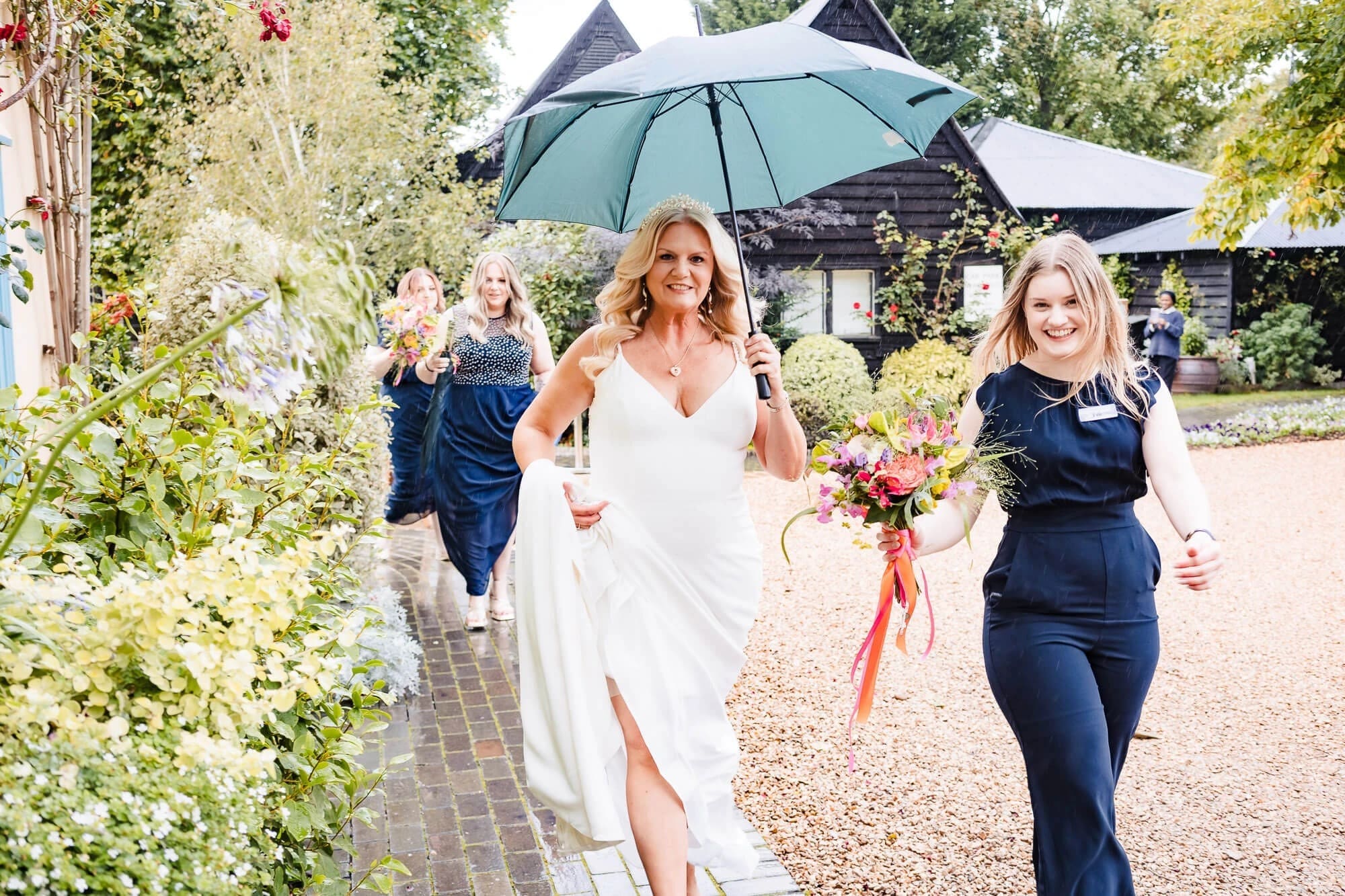 Bride walking down a gravel path with bridesmaids, holding an umbrella and bouquet. — Wedding at South Farm, Cambridgeshire