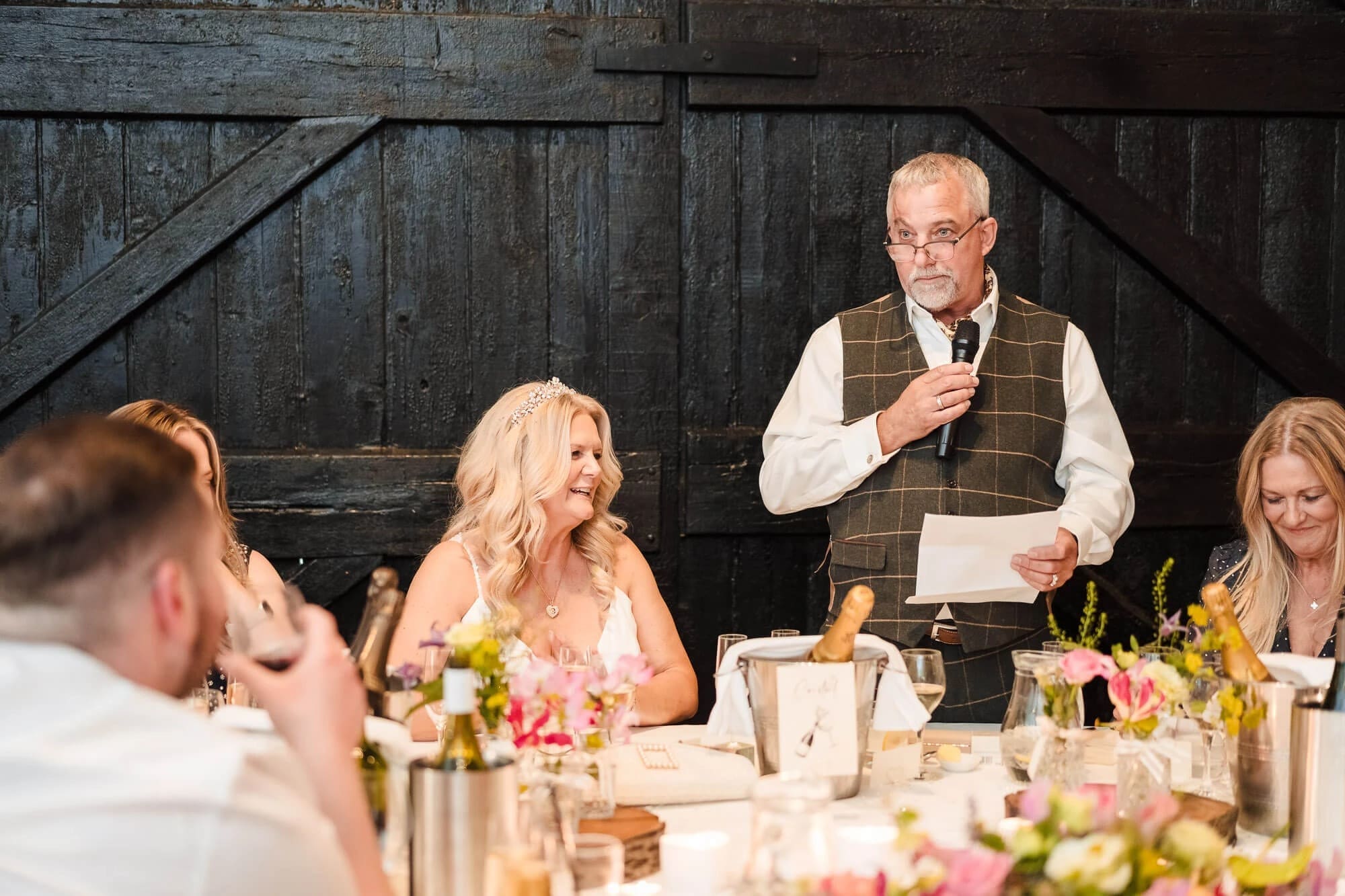 Man giving a speech at a wedding reception dinner, gesturing while speaking to seated guests. — Wedding at South Farm, Cambridgeshire