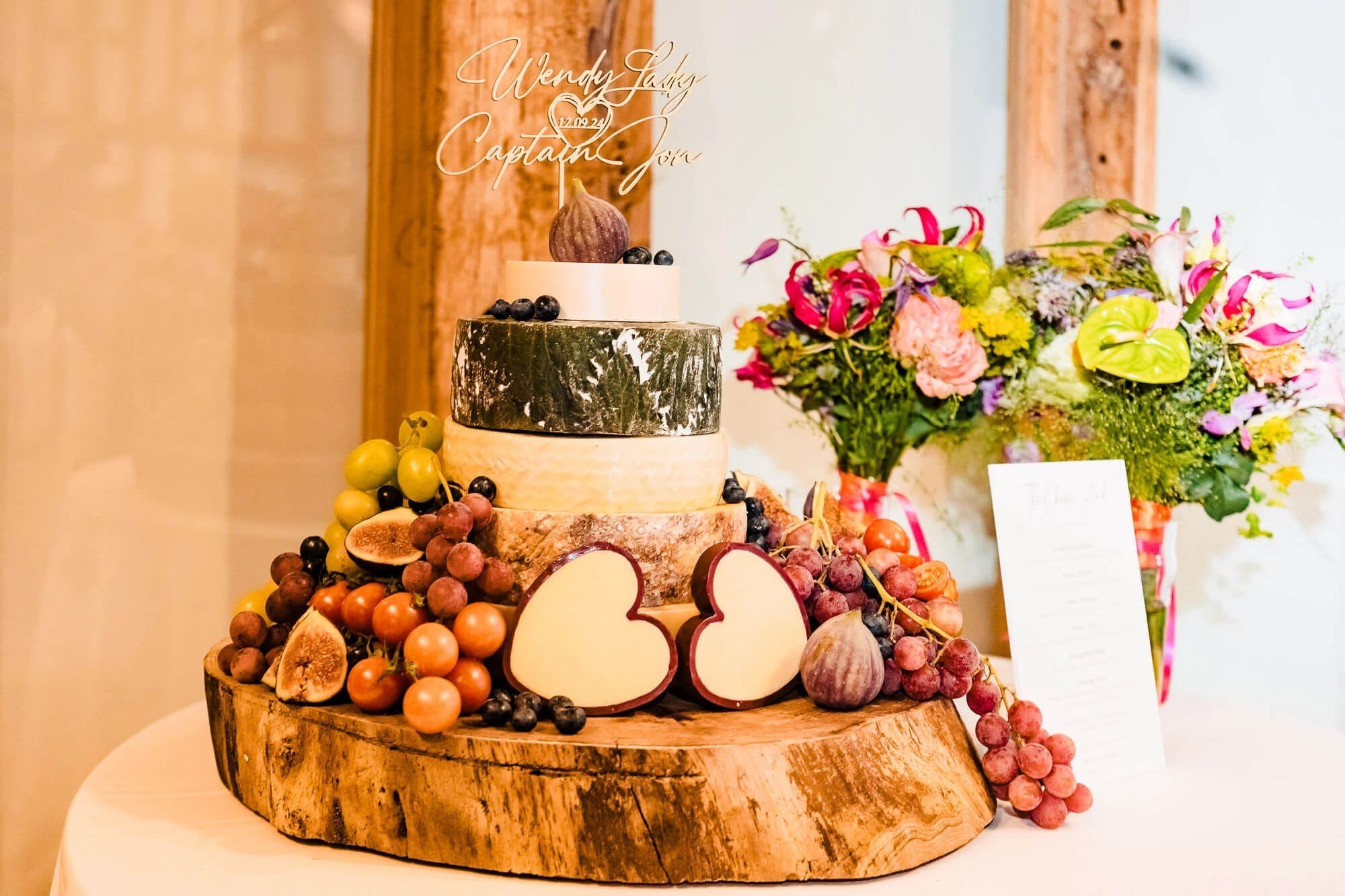 A tiered wedding cake displayed on a wooden base surrounded by fresh fruit and flowers. — Wedding at South Farm, Cambridgeshire