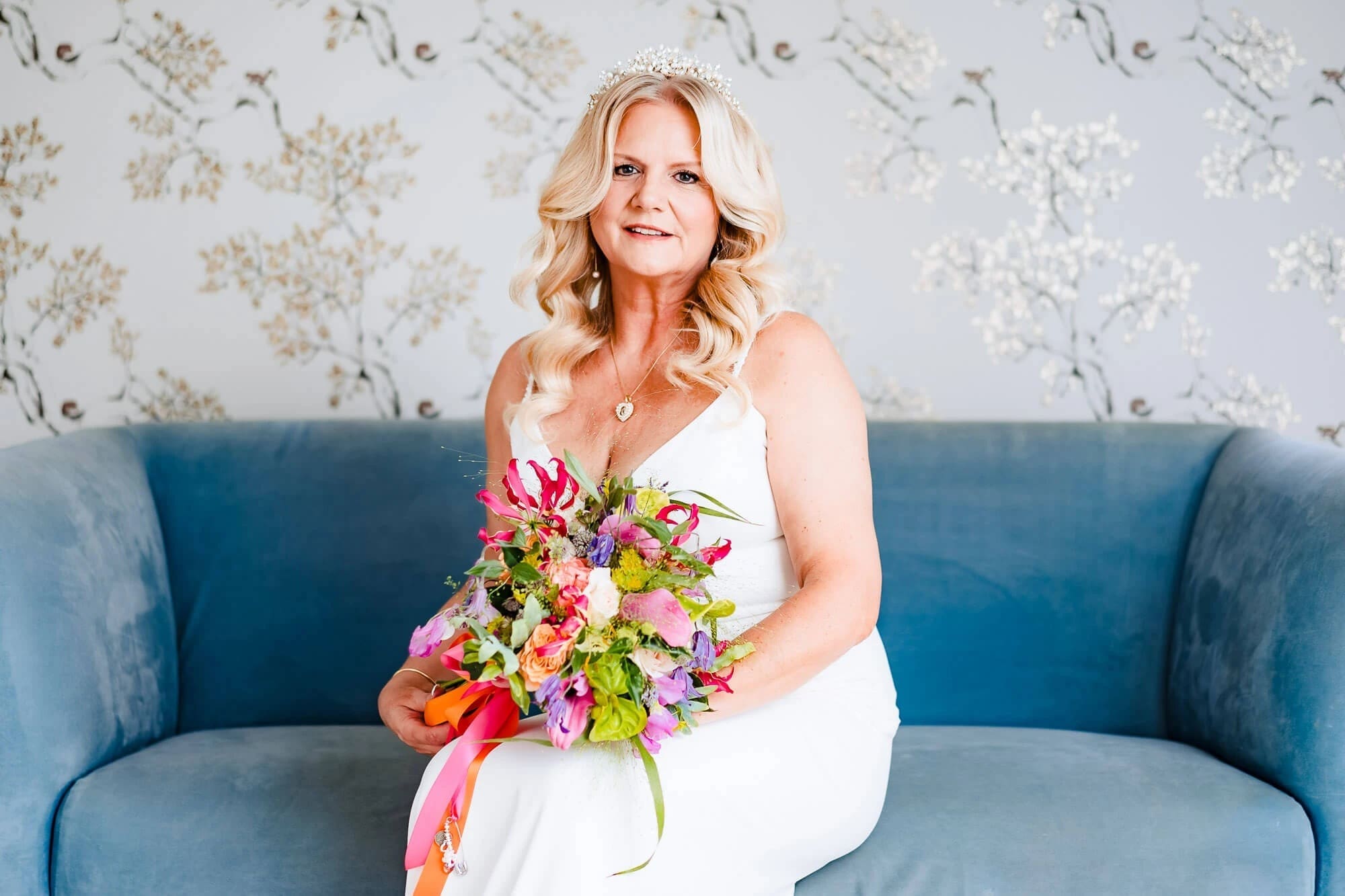 Woman in white dress sitting on blue sofa holding a colorful bouquet, smiling at camera. — Wedding at South Farm, Cambridgeshire