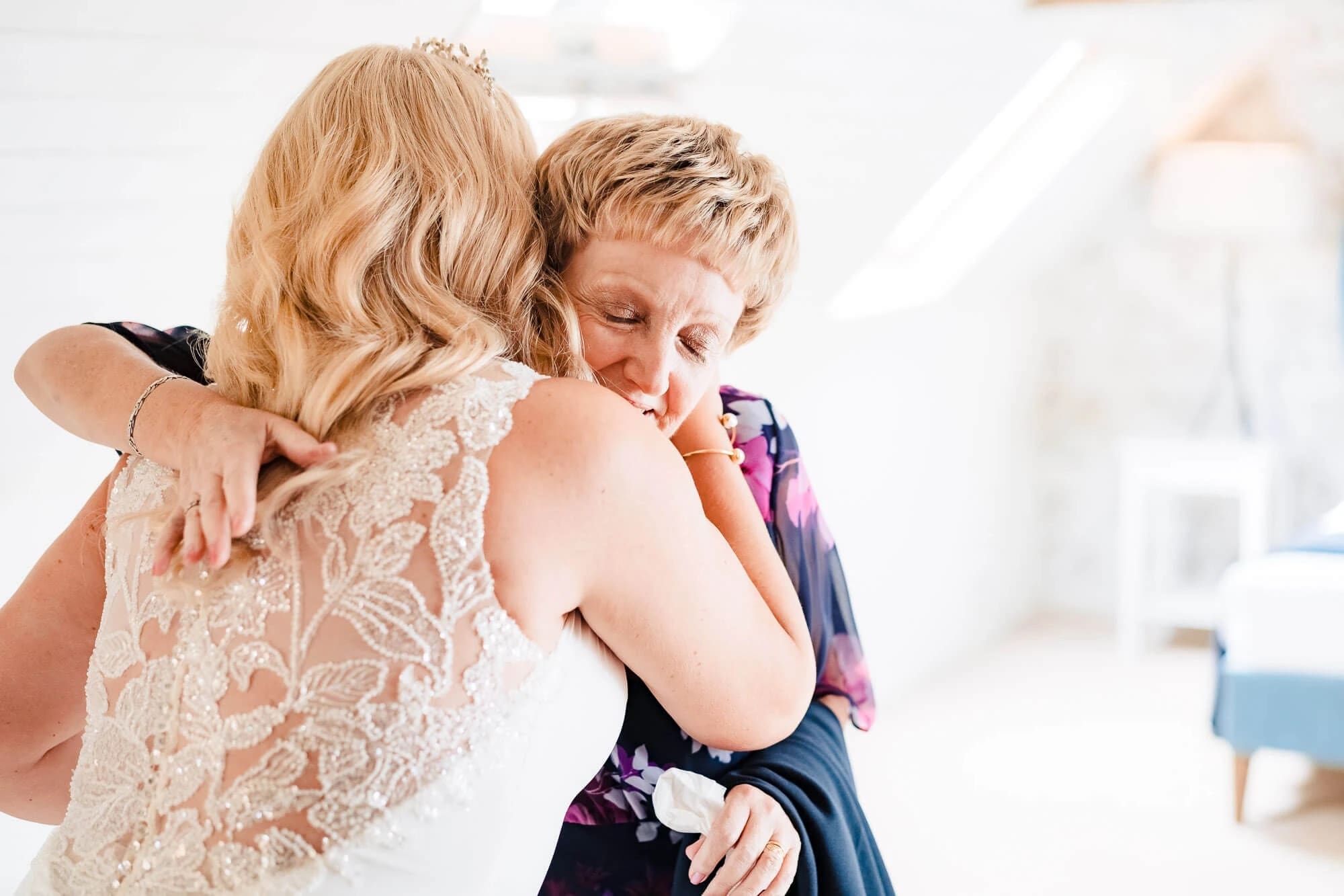 Young child hugging a woman in a lace dress from behind, both smiling with eyes closed. — Wedding at South Farm, Cambridgeshire