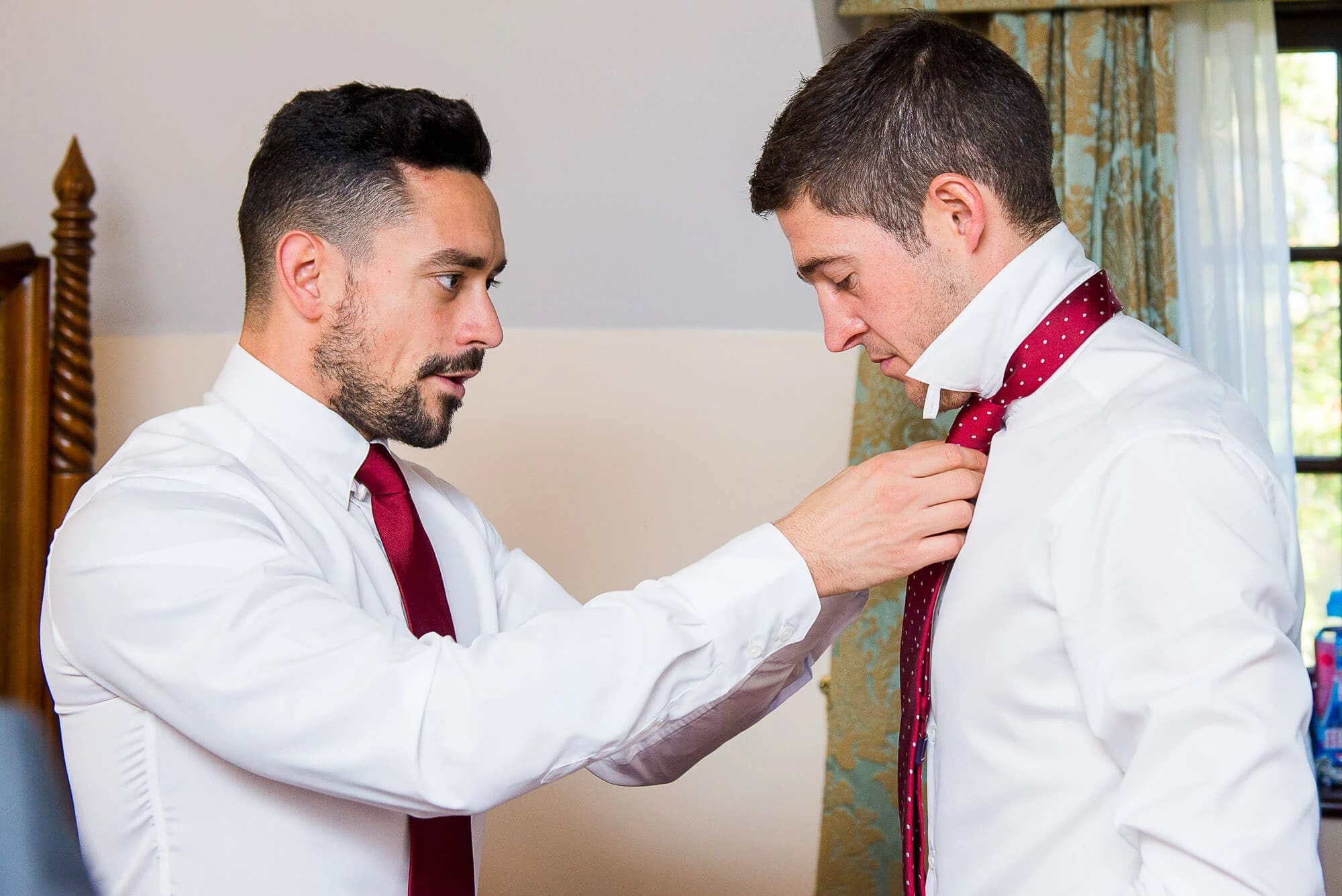 Groomsman adjusting another man's burgundy tie before the ceremony. — Wedding at Stanhill Court Hotel, Surrey