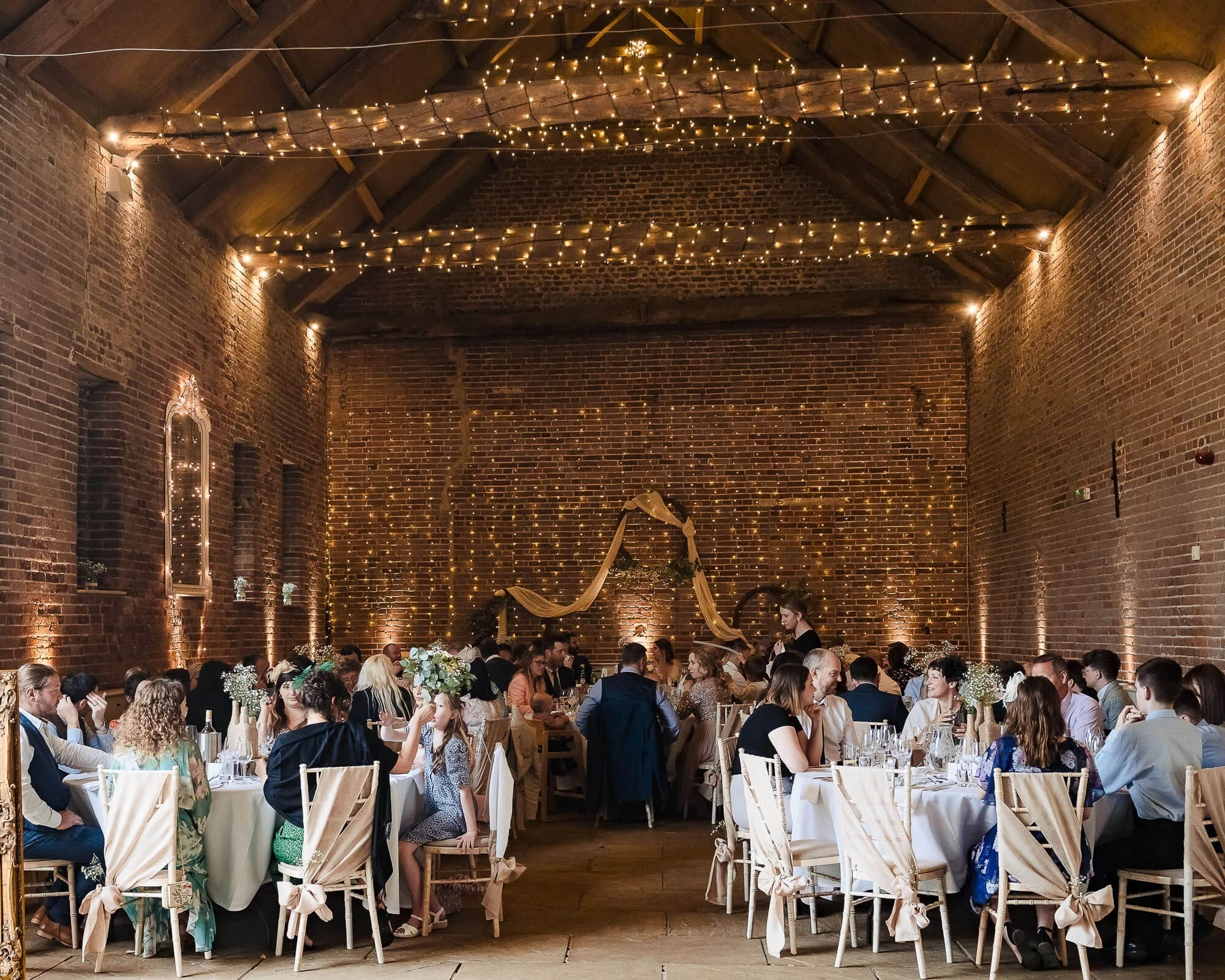 Guests dining at long tables during a wedding reception dinner in a decorated brick venue. — Wedding at Manor Mews, Norfolk