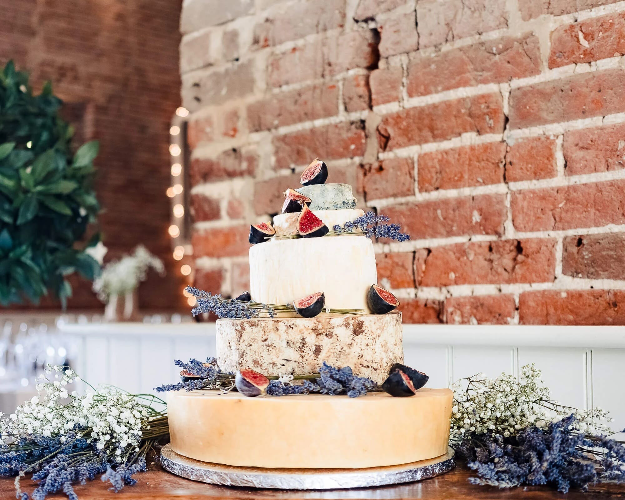 A three-tiered wedding cake decorated with lavender sprigs and fabric bird toppers sits displayed against a brick wall. — Wedding at Manor Mews, Norfolk