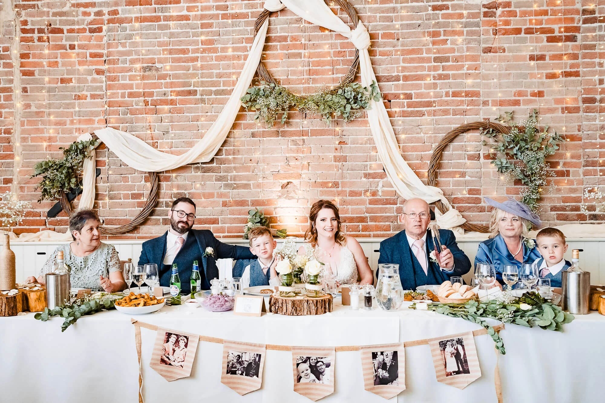 Wedding party seated at head table during reception dinner, with family photos displayed on banner below. — Wedding at Manor Mews, Norfolk