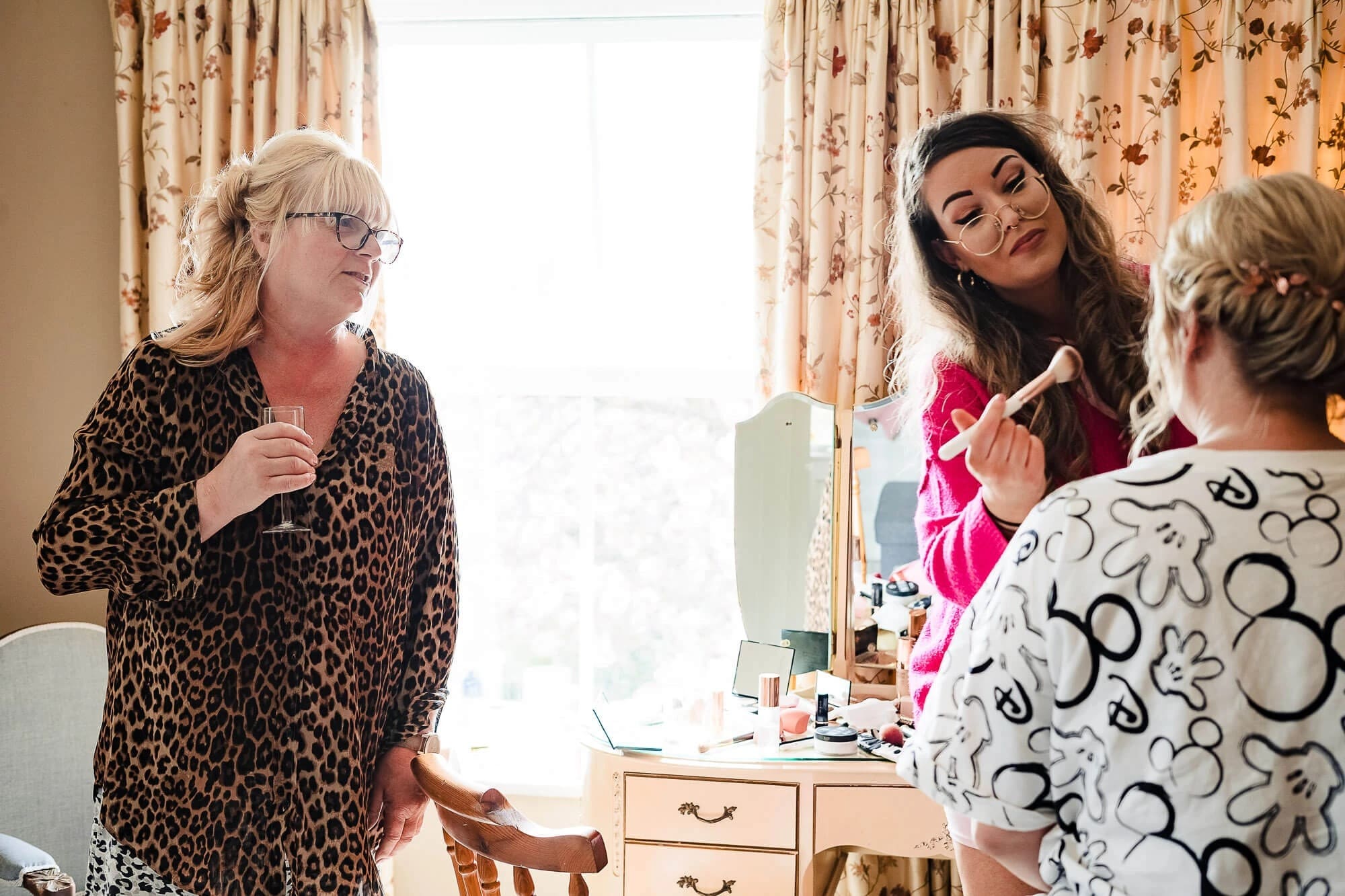 Woman applying makeup while two others watch and assist in a bedroom mirror setup. — Wedding at Manor Mews, Norfolk