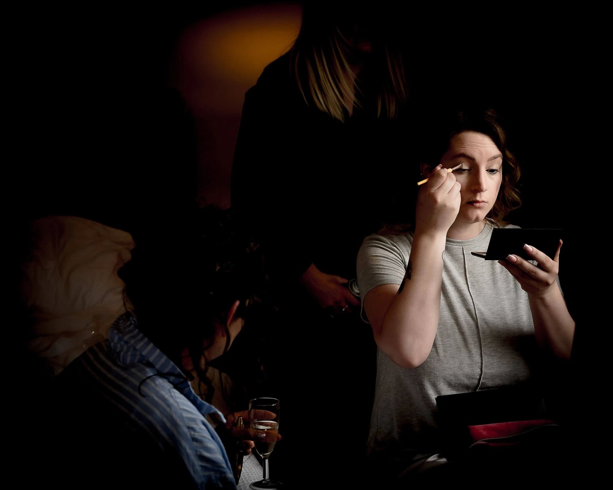 Woman applying makeup while being assisted by another person in a darkened space. — Wedding at Manor Mews, Norfolk