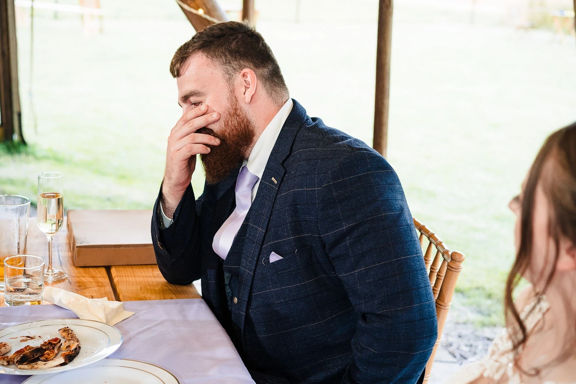 Bearded man in navy suit pauses thoughtfully during a meal, hand to face, at an outdoor dining table. — Wedding at Moulton Parish Church, Northamptonshire