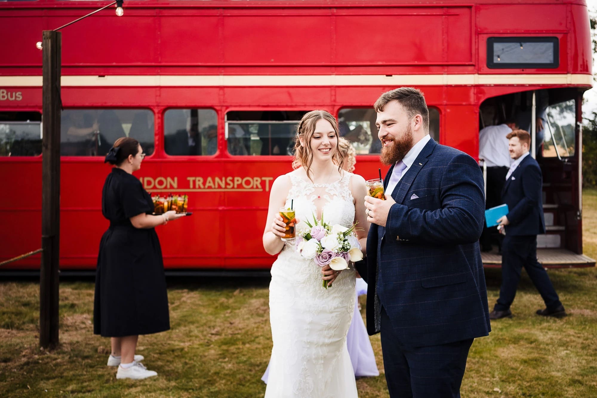 Bride and groom holding drinks and smiling together outside a red double-decker bus at their wedding reception. — Wedding at Moulton Parish Church, Northamptonshire