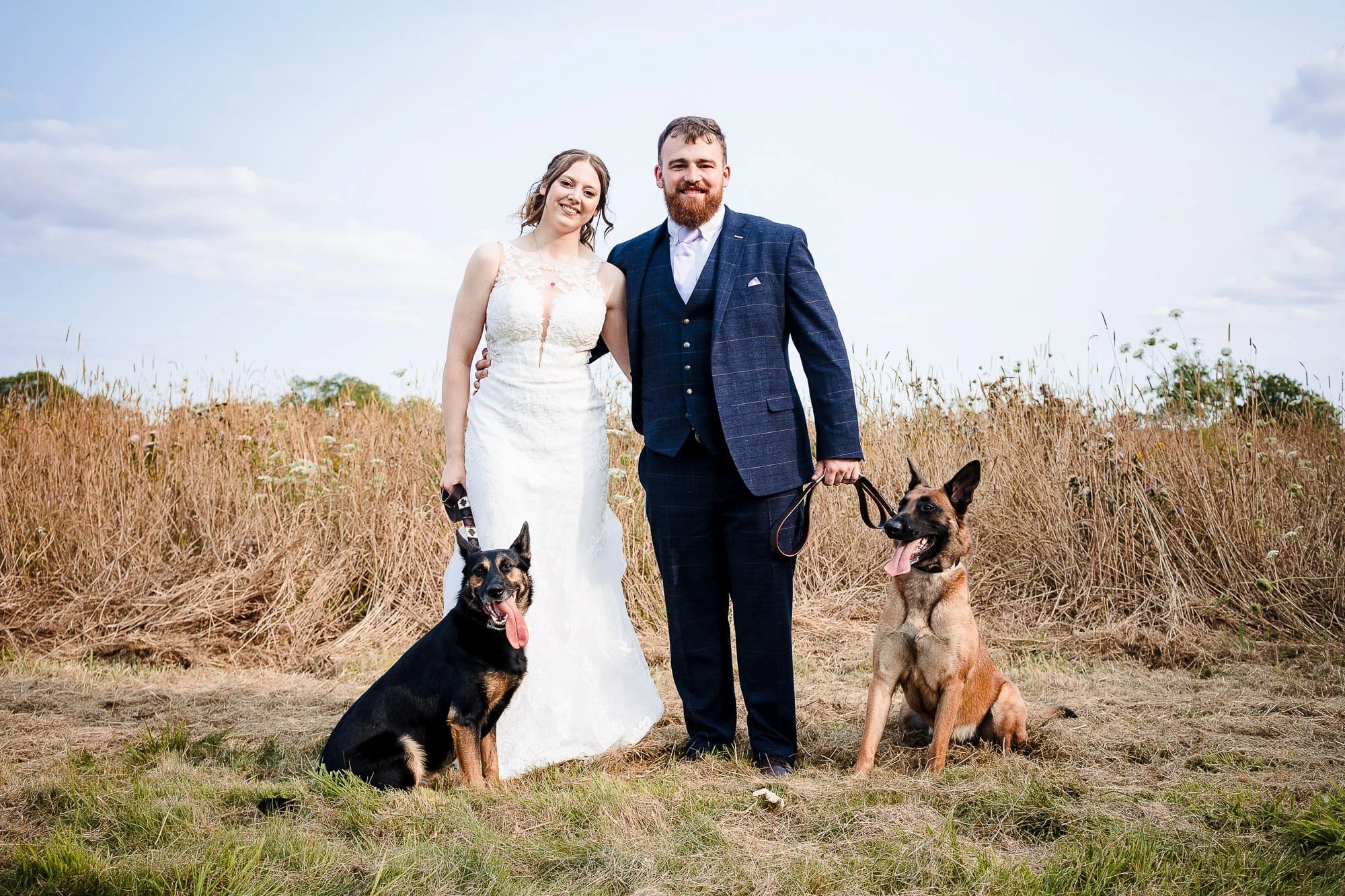 Bride and groom stand together in an open field with their two dogs, posing for a formal wedding portrait. — Wedding at Moulton Parish Church, Northamptonshire