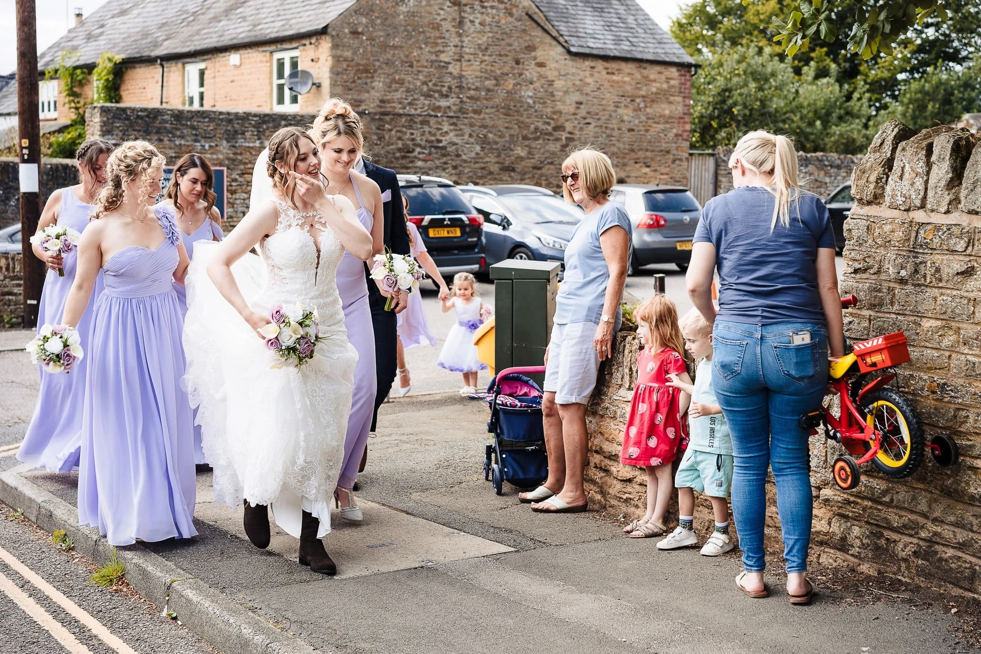 Bride walking down a village street with bridesmaids and wedding guests, including children with toys. — Wedding at Moulton Parish Church, Northamptonshire