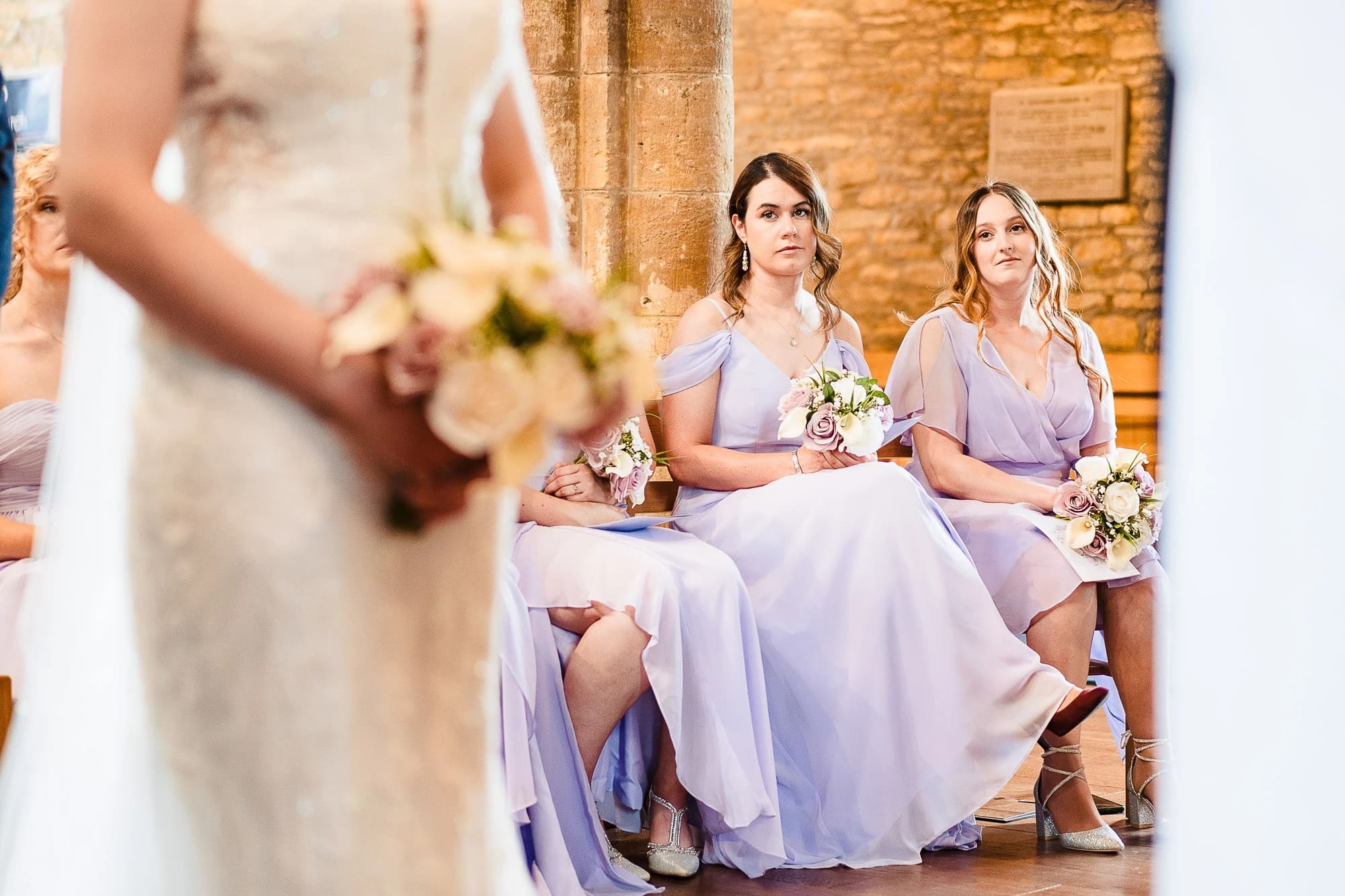 Bridesmaids in lavender dresses sit holding bouquets, watching the bride prepare in the foreground. — Wedding at Moulton Parish Church, Northamptonshire