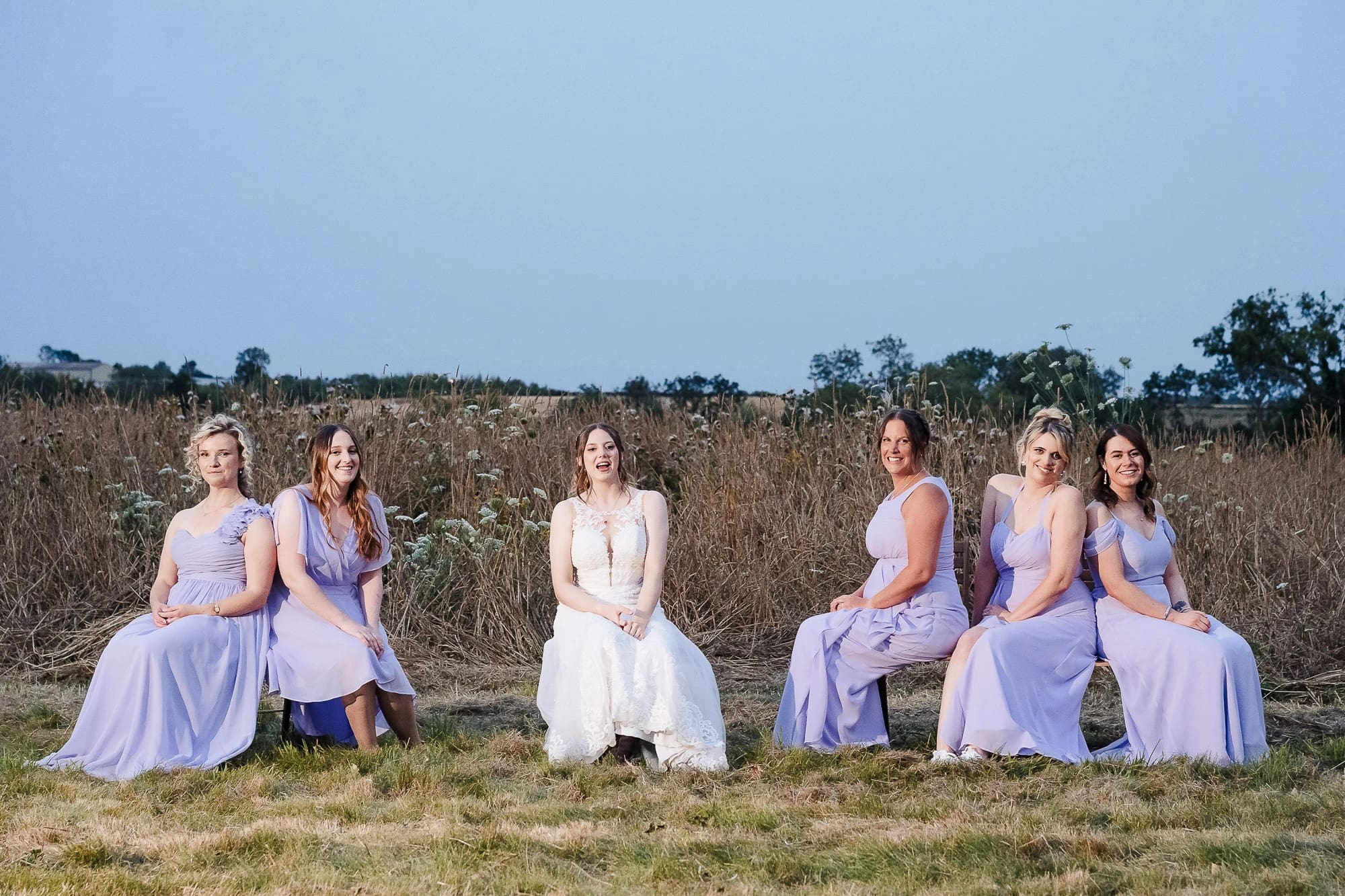 Six women in bridesmaid dresses seated and standing in a line across a dried field for a formal portrait. — Wedding at Moulton Parish Church, Northamptonshire