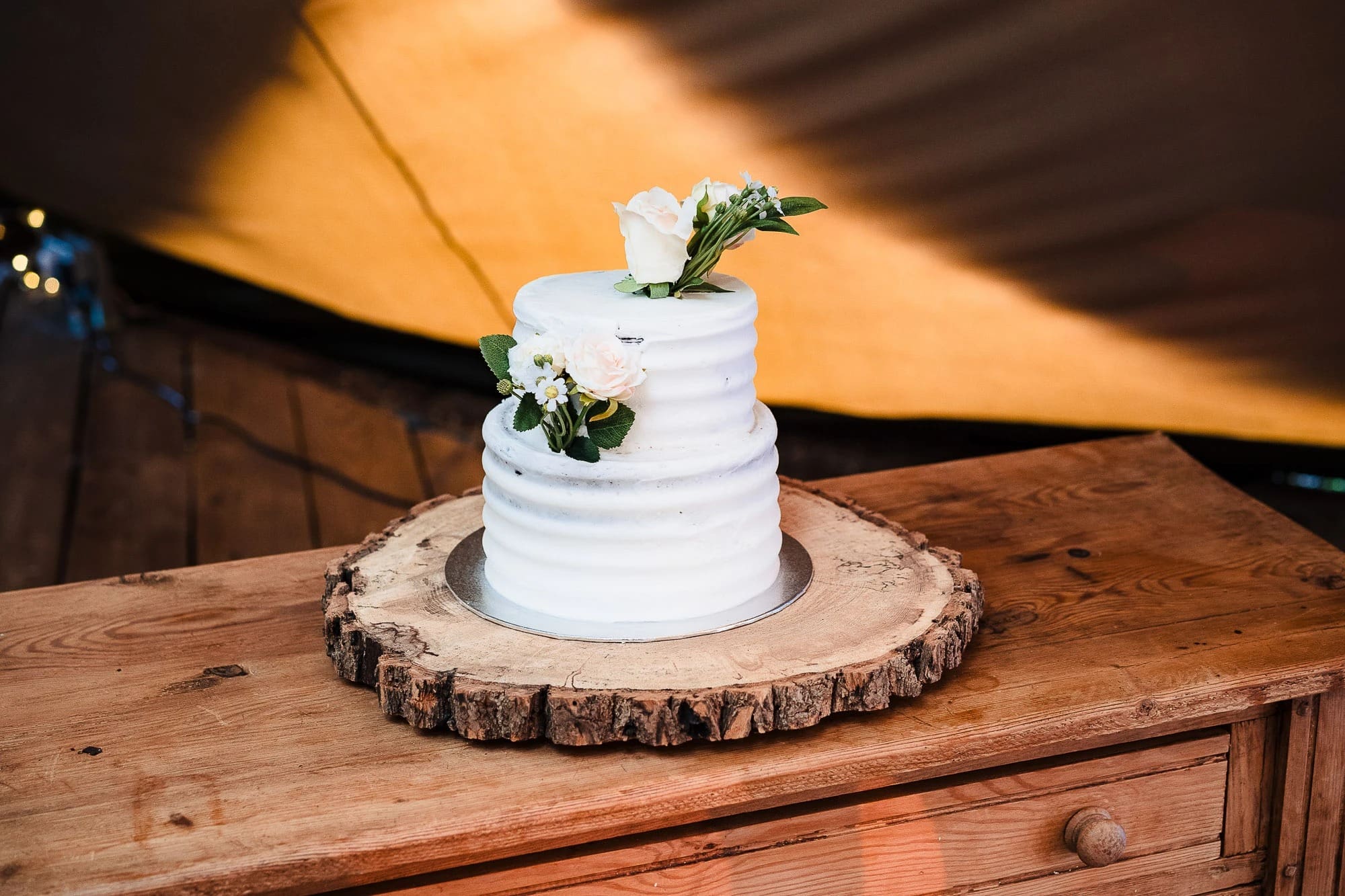 A white tiered wedding cake sits displayed on a wooden slice, decorated with fresh white flowers and greenery. — Wedding at Moulton Parish Church, Northamptonshire