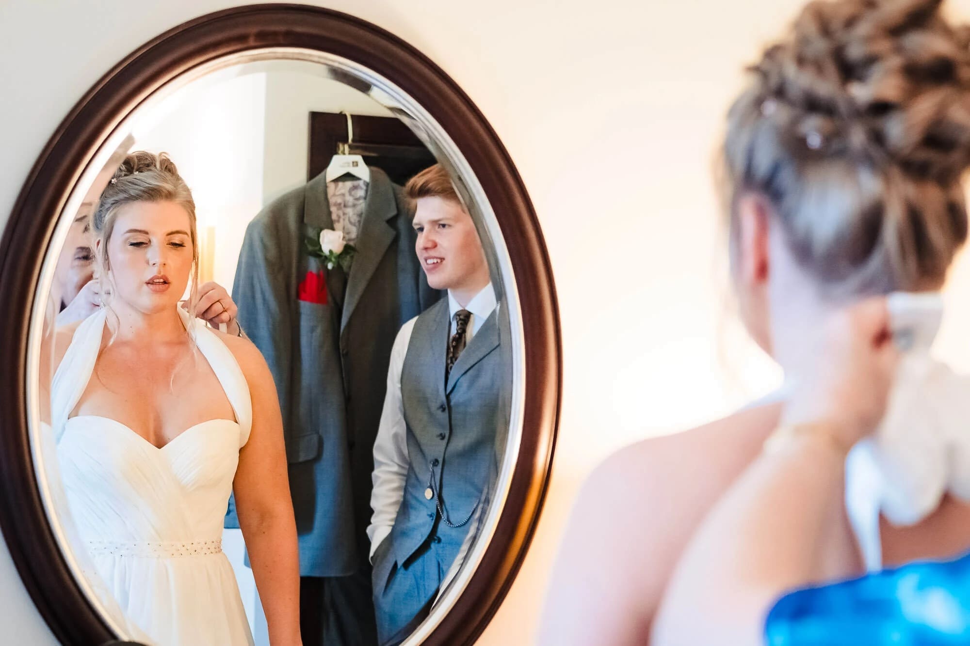 Bride admiring herself in an ornate mirror while wedding guests and family members stand behind her in reflection. — Wedding at Sprowston Manor Hotel, Norfolk
