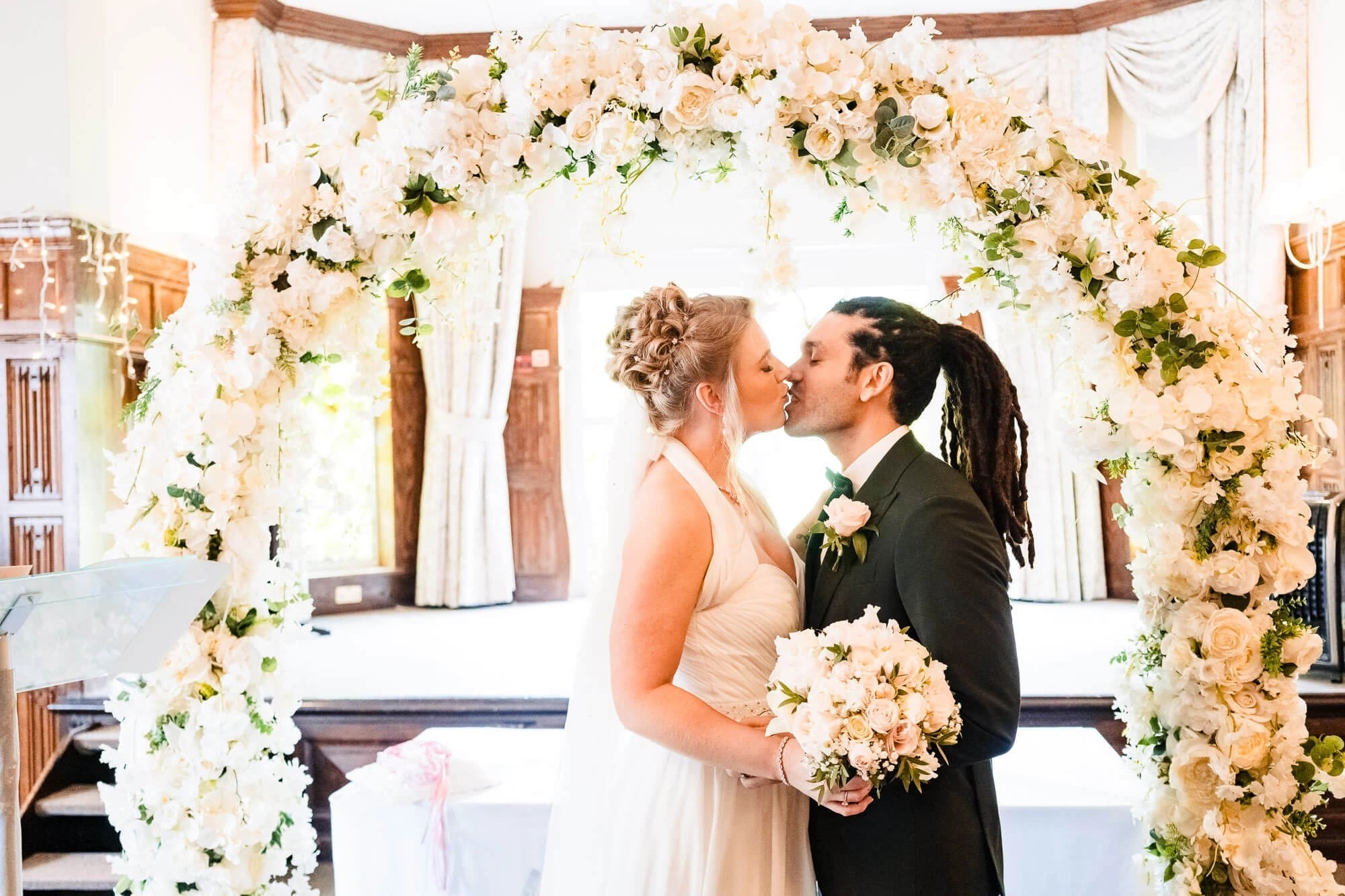 Bride and groom kiss under a floral arch during their wedding ceremony. — Wedding at Sprowston Manor Hotel, Norfolk