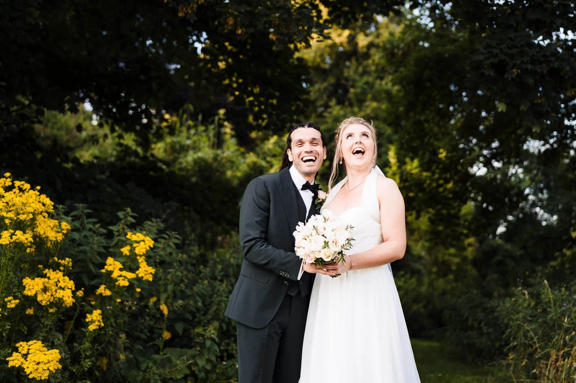 Bride and groom laughing together outdoors, holding her bouquet, both looking upward with joy. — Wedding at Sprowston Manor Hotel, Norfolk