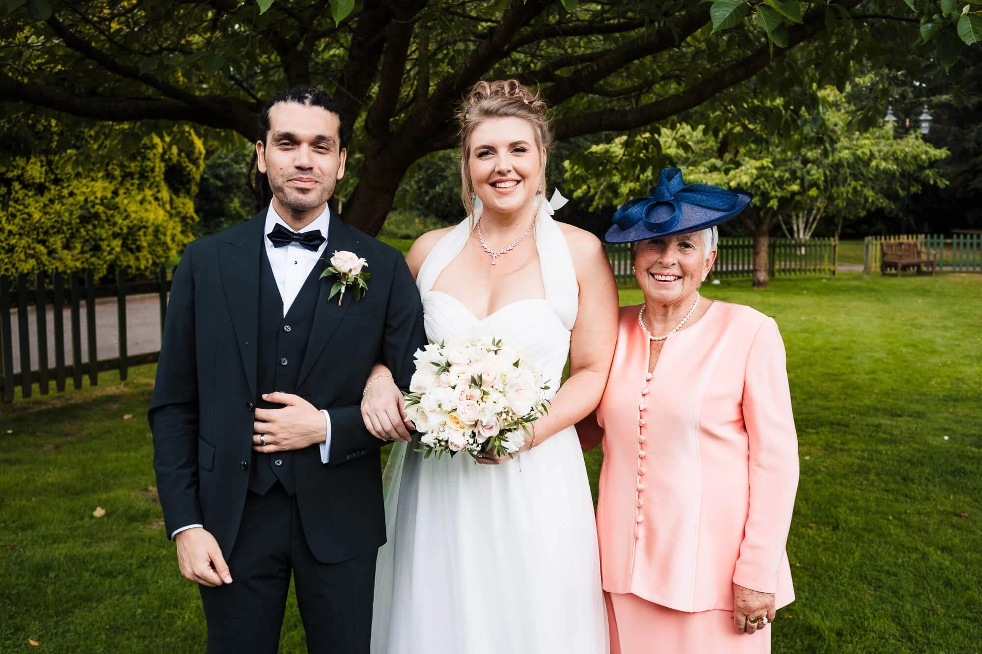Bride posing with groom and mother for a formal family portrait on the lawn. — Wedding at Sprowston Manor Hotel, Norfolk
