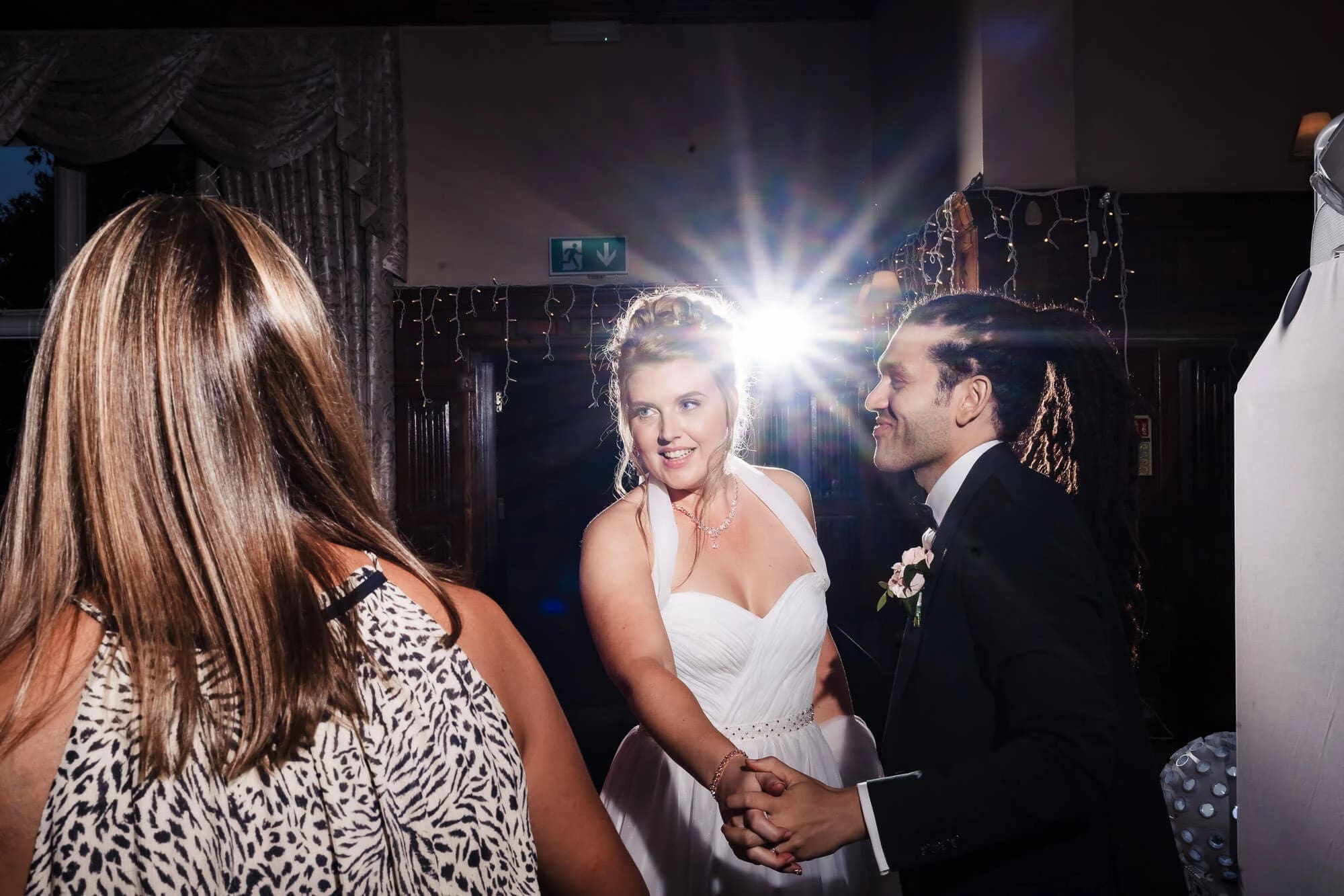 Woman in white dress conversing with man in dark suit at an indoor event, bright flash photography lighting the interaction. — Wedding at Sprowston Manor Hotel, Norfolk